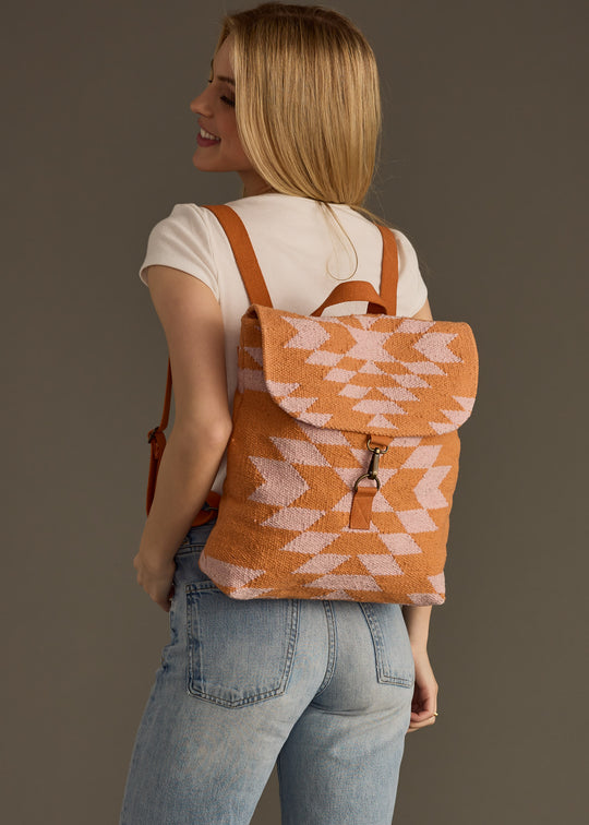 A smiling woman with long blonde hair wears a white t-shirt and light blue jeans, posing with the panacheaccessories Elisa Backpack, which features adjustable straps and a vibrant orange pattern, slung over her shoulder against a plain background.
