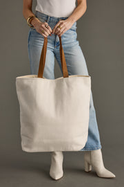 A person in blue jeans, a white shirt, and white boots is holding the Skiing People Tote from Panache Apparel Collection, featuring leather suede straps, while standing against a gray background.