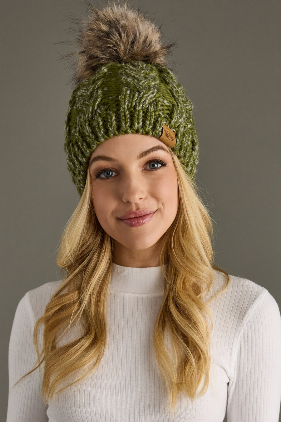Woman wearing a green knitted beanie with a fur pom-pom against a gray background