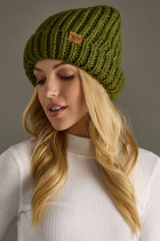 Woman wearing a green knitted beanie with a brand logo against a gray background
