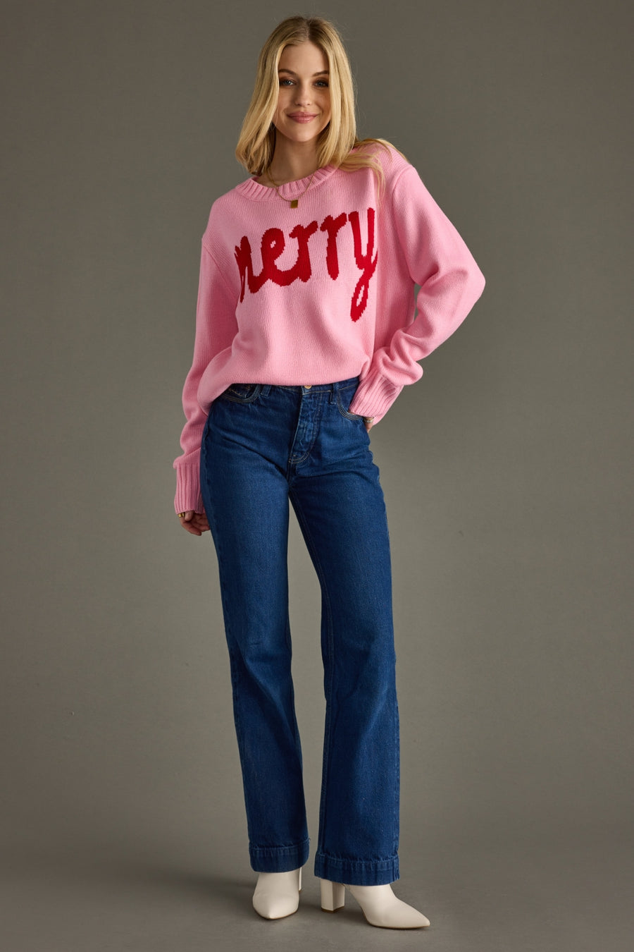 Woman wearing a pink sweater with 'Merry' text and blue jeans on a gray background