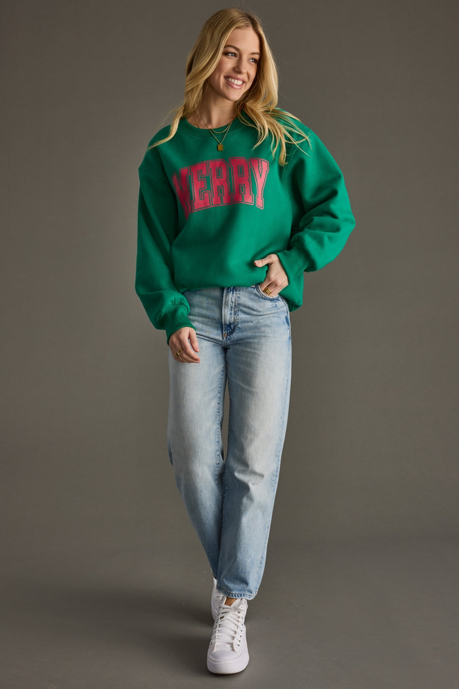 Woman wearing a green sweater with 'Merry' text, light blue jeans, and white sneakers on a gray background