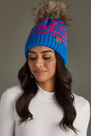 Woman wearing a blue and pink beanie with a fur pom-pom on a gray background