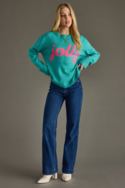 Woman wearing a teal sweater with 'jolly' in pink text and blue jeans on a gray background