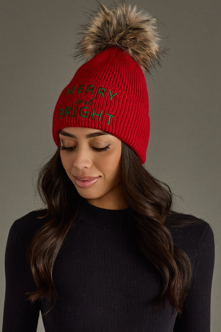 A woman with long dark hair wears the Panache Apparel Collection Merry & Bright Pom Hat - Red & Green, paired with a black ribbed top. She looks down slightly with a soft smile.