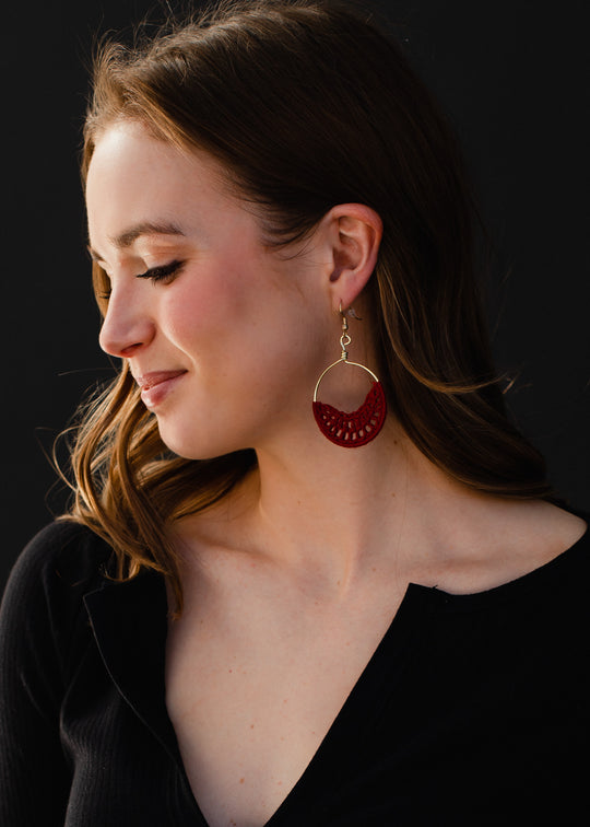 A woman with light brown hair looks down and smiles softly, wearing a black top and striking Sadie Crochet Earrings in burgundy by panacheaccessories, set against a plain dark background.