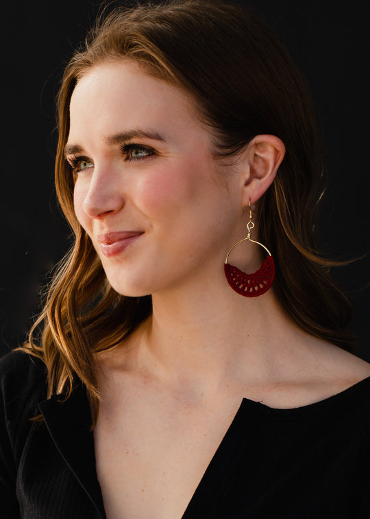 A young woman with wavy brown hair smiles softly to the side against a dark background, wearing panacheaccessories large Sadie Crochet Earrings in burgundy and a black top.