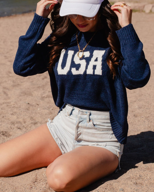 Woman sitting on sand, wearing a navy blue USA sweater, light denim shorts, a white baseball cap, sunglasses, and a gold necklace. Her face is partially covered by the cap.