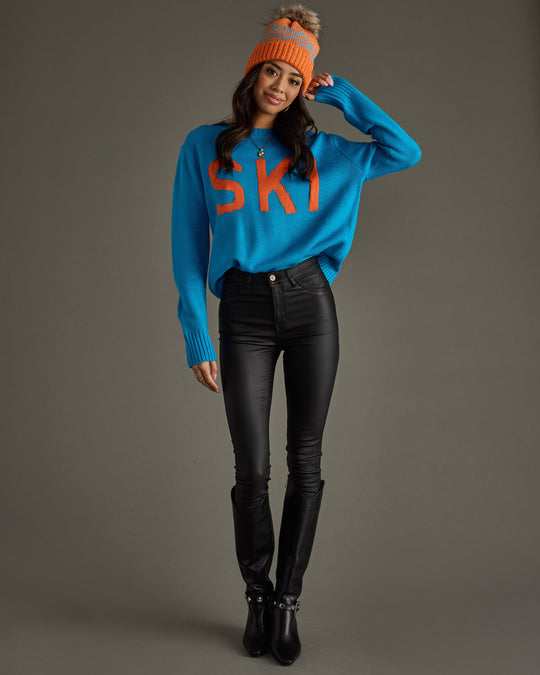 A woman wearing a blue sweater with SKI in orange letters, black pants, black boots, and an orange knit beanie with a pom-pom stands against a plain gray background.