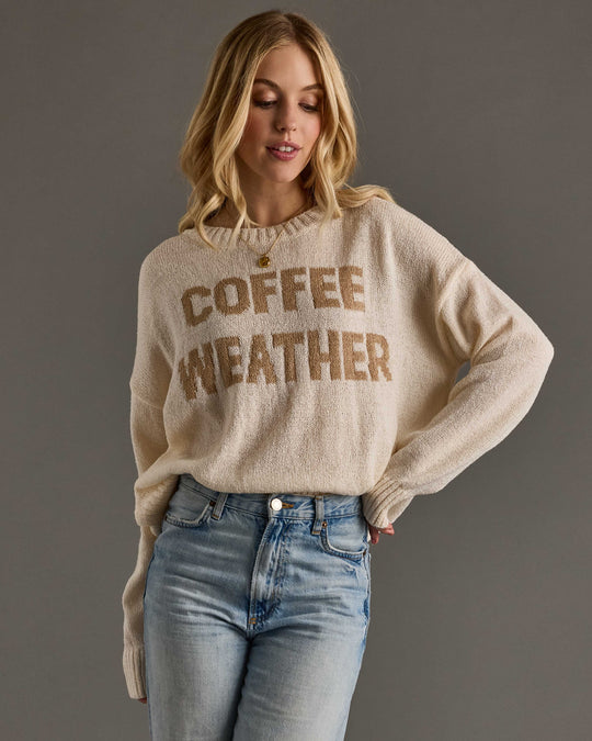 A woman with long blonde hair wearing a beige sweater that says COFFEE WEATHER and light blue jeans stands against a plain gray background.