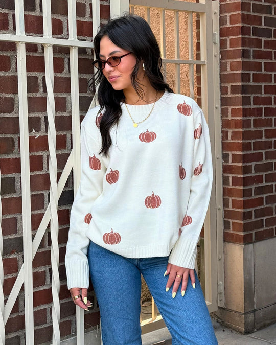 Woman wearing sunglasses, a white sweater with pumpkin prints, blue jeans, and yellow nails stands by a brick wall and metal gate, looking to the side.