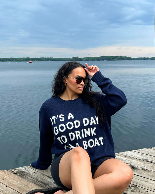 A woman in sunglasses sits on a dock by a lake, wearing a navy sweater that says ITS A GOOD DAY TO DRINK ON A BOAT. The sky is cloudy, and the water and distant shoreline are visible.