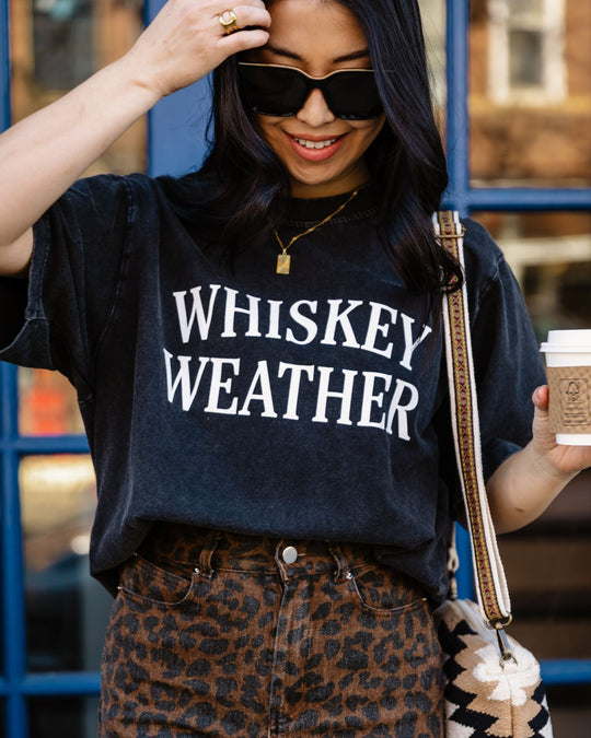 Woman wearing sunglasses and a black t-shirt that says WHISKEY WEATHER, paired with a leopard print skirt, holding a coffee cup and touching her hair, standing in front of a blue door.