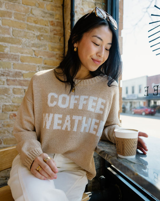 A woman with long dark hair sits by a window in a café, holding a cup of coffee and wearing a tan sweater that says COFFEE WEATHER. She is looking down and smiling softly.