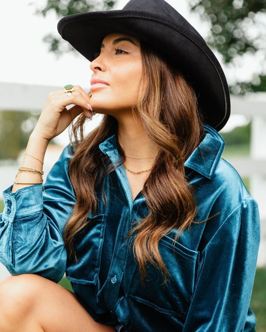 A woman wearing a black cowboy hat and a teal long-sleeved shirt with her hand resting on her chin.