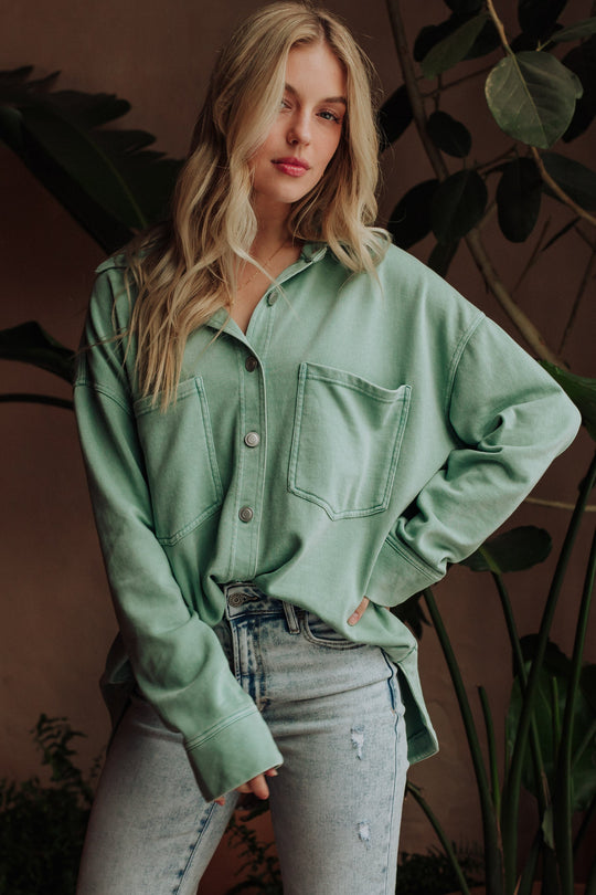 A woman with long blonde hair wearing a mauve button-up shirt and light blue jeans stands in front of green indoor plants, looking down with one hand in her pocket.