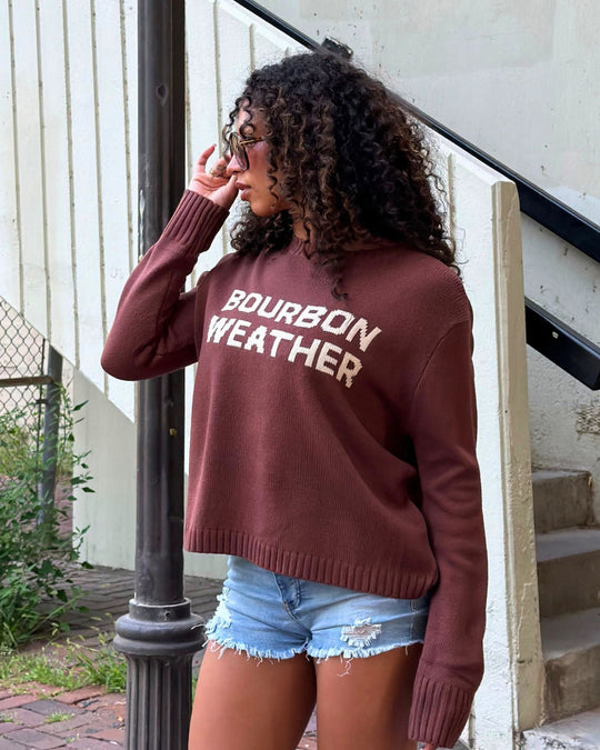 Woman with curly hair wearing sunglasses, a brown sweater that says BOURBON WEATHER, and denim shorts stands outdoors near stairs and a pole, looking to the side.