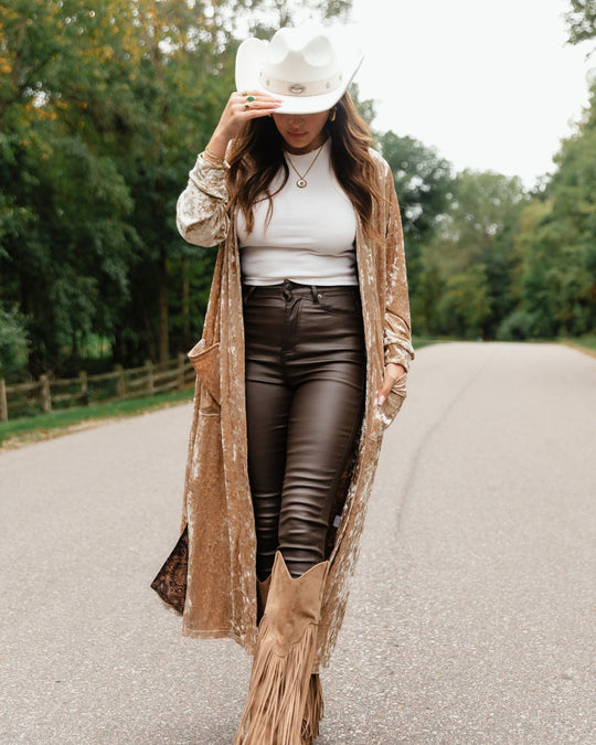 A woman wearing a white cowboy hat, white top, brown pants, tan fringed boots, and a long beige duster walks down a paved road.