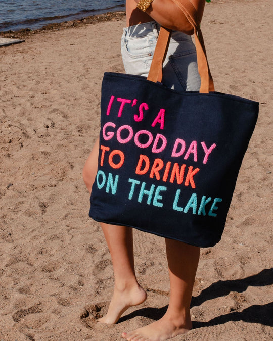 A person holding a navy tote bag with colorful text that reads, ITS A GOOD DAY TO DRINK ON THE LAKE, standing barefoot on a sandy beach near the water.