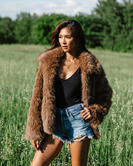 Woman in a brown fur coat, black top, and denim shorts stands in a grassy field with trees in the background, looking to the side under a sunny sky.