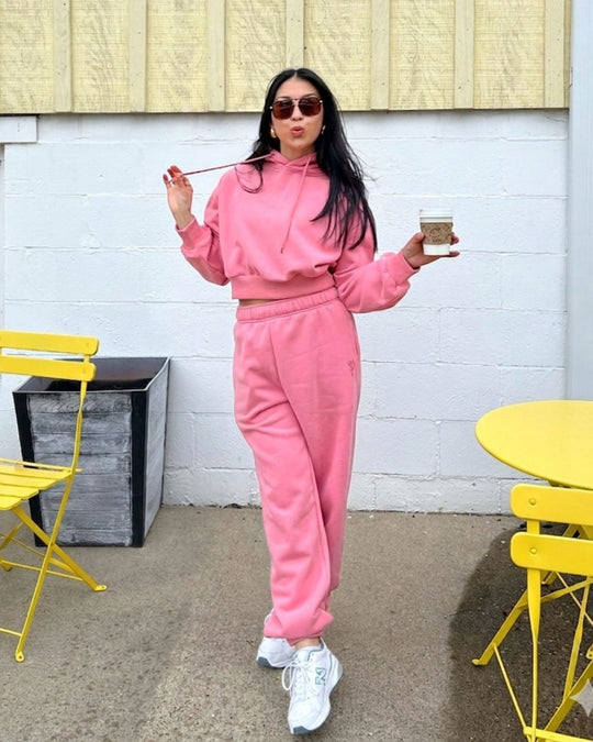 A woman with long black hair smiles at the camera, wearing a bright green hoodie and matching sweatpants. She stands indoors against a plain white wall.