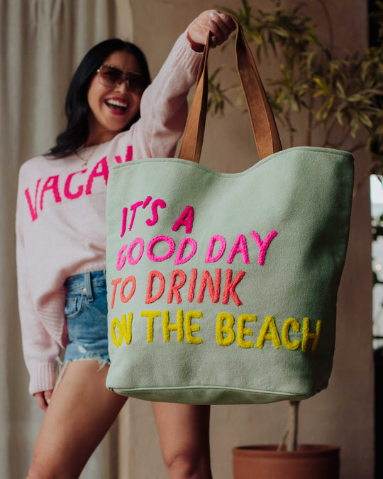 A smiling woman in sunglasses and a VACAY sweater holds a large green tote bag reading, Its a good day to drink on the beach, in colorful letters.