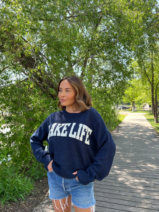 A person holding a navy tote bag with colorful text that reads, ITS A GOOD DAY TO DRINK ON THE LAKE, standing barefoot on a sandy beach near the water.