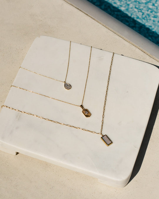 Three delicate gold necklaces with small geometric pendants are displayed on a white marble slab near a swimming pool, with sunlight casting soft shadows.