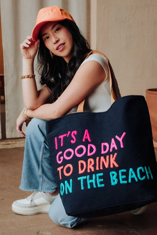 A woman in a peach cap and casual clothes sits indoors, holding a navy tote bag with colorful text that reads, Its a good day to drink on the beach.