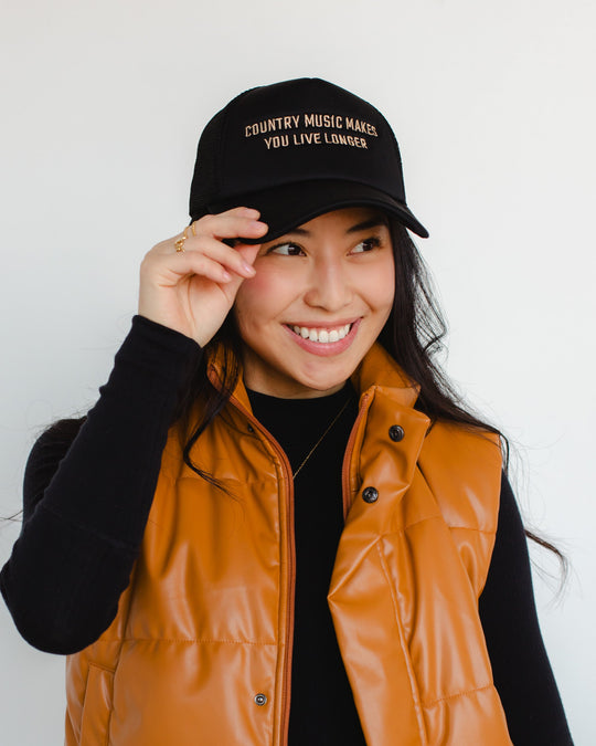 A smiling woman in a black long-sleeve shirt and brown vest tilts her black hat that reads Country Music Makes You Live Longer against a plain white background.