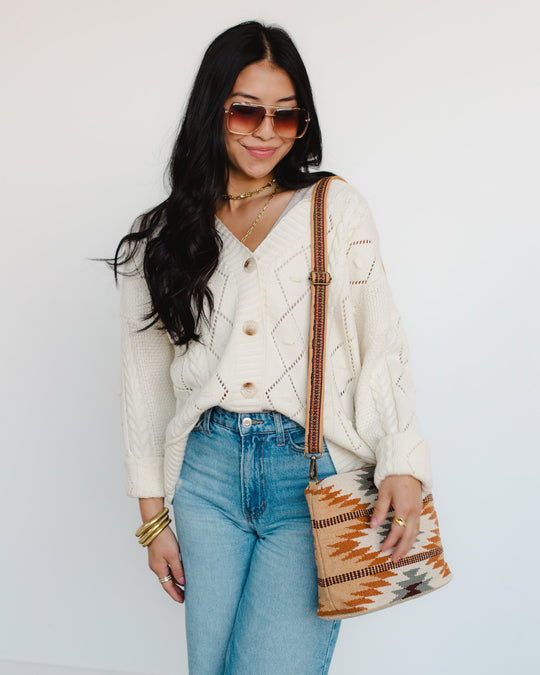 Woman with long dark hair wearing sunglasses, a cream knit cardigan, blue jeans, and gold bracelets, carrying a patterned shoulder bag with neutral and brown tones, standing against a white background.
