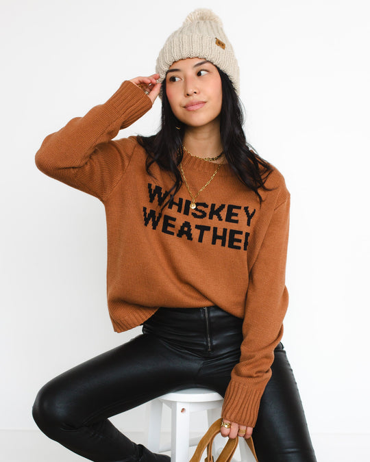 Woman wearing a beige knit beanie, brown sweater with WHISKEY WEATHER text, black leather pants, and gold necklace sits on a stool, looking to the side with one hand touching her hat.