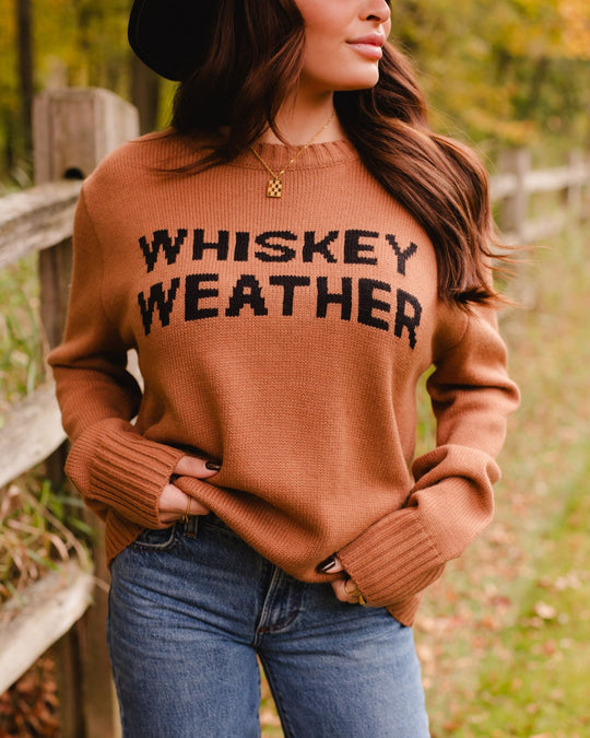 A woman wearing a brown sweater that says WHISKEY WEATHER in black lettering.