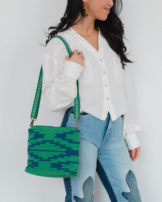 A woman wearing a white blouse and high-waisted jeans with heart patches holds a green and blue patterned shoulder bag with a decorative strap. She has long dark hair and gold hoop earrings.