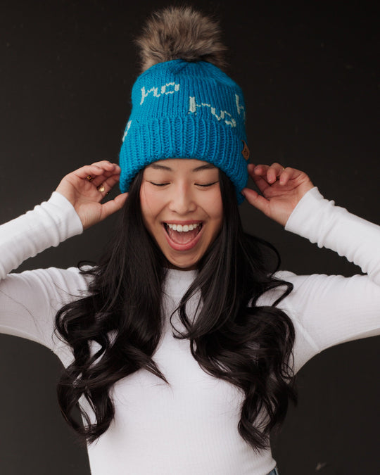 A smiling woman with long black hair wears a blue knit hat with a pom-pom and a white long-sleeve shirt, holding the sides of her hat against a dark background.