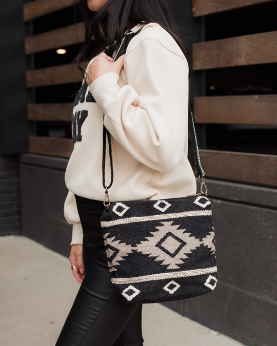 Woman wearing a cream sweatshirt and black pants, holding a black and cream geometric-patterned shoulder bag, standing outside near a modern wooden slat wall.