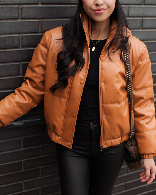 Woman wearing a brown faux leather puffer jacket, black top, and black pants, standing against a dark brick wall and holding a purse with a chain strap.