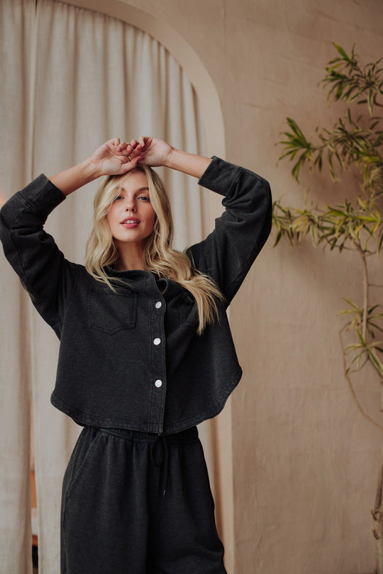 A woman with long blonde hair wears a black button-up shirt over a white crop top and black pants, standing indoors in front of beige curtains and a potted plant.