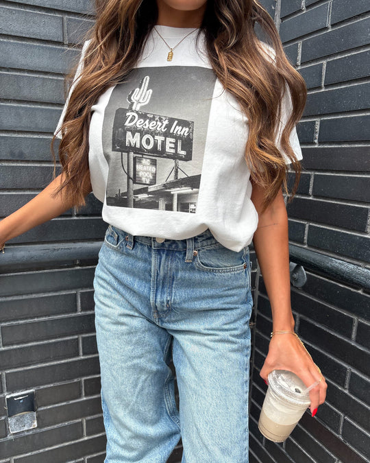 A woman in a white graphic Desert Inn Motel T-shirt and light blue jeans stands against a dark brick wall, holding an iced coffee. Her long wavy hair is loose and she wears a gold necklace.