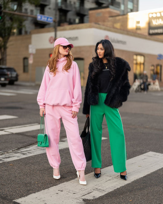 Two women standing in a crosswalk; one wears a pink sweatsuit and cap with a green purse, and the other wears a black fur jacket and green trousers.