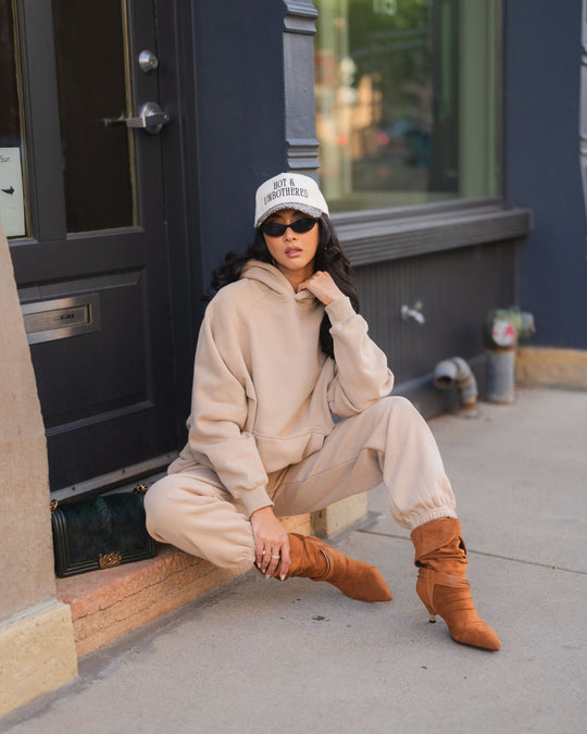 A person sitting on a doorstep wearing a beige hoodie and matching sweatpants, a baseball cap that reads HOT & UNBOTHERED, brown heeled boots, and black sunglasses, with a small handbag beside them.