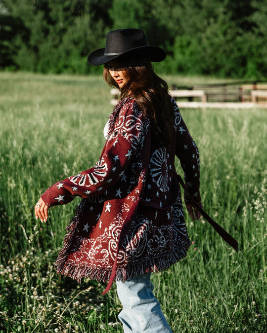 A person in a black hat, maroon patterned cardigan, and jeans walks through tall grass outdoors with trees in the background on a sunny day.