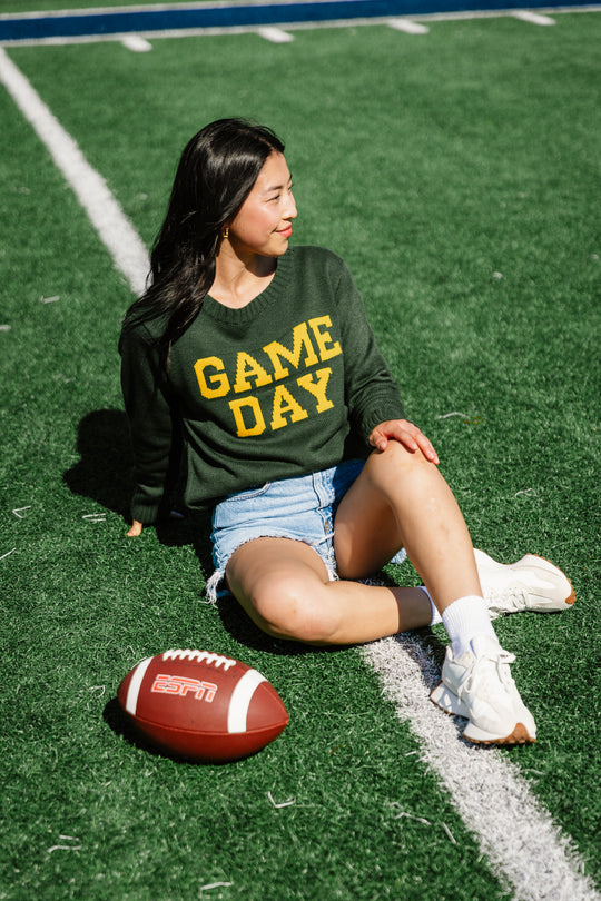 A woman sits on a football field smiling, wearing a green Game Day sweater, denim shorts, and white sneakers, with a football labeled ESPN resting by her legs.