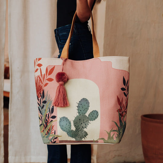 A person holds a large canvas tote bag with tan handles, featuring a colorful cactus and foliage design, a pink background, and a decorative pink tassel. The person is wearing dark jeans and a black top.