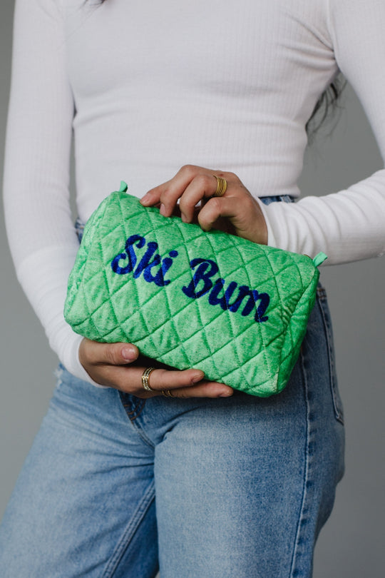 Person holding a green quilted pouch with 'Ski Bum' text against a gray background