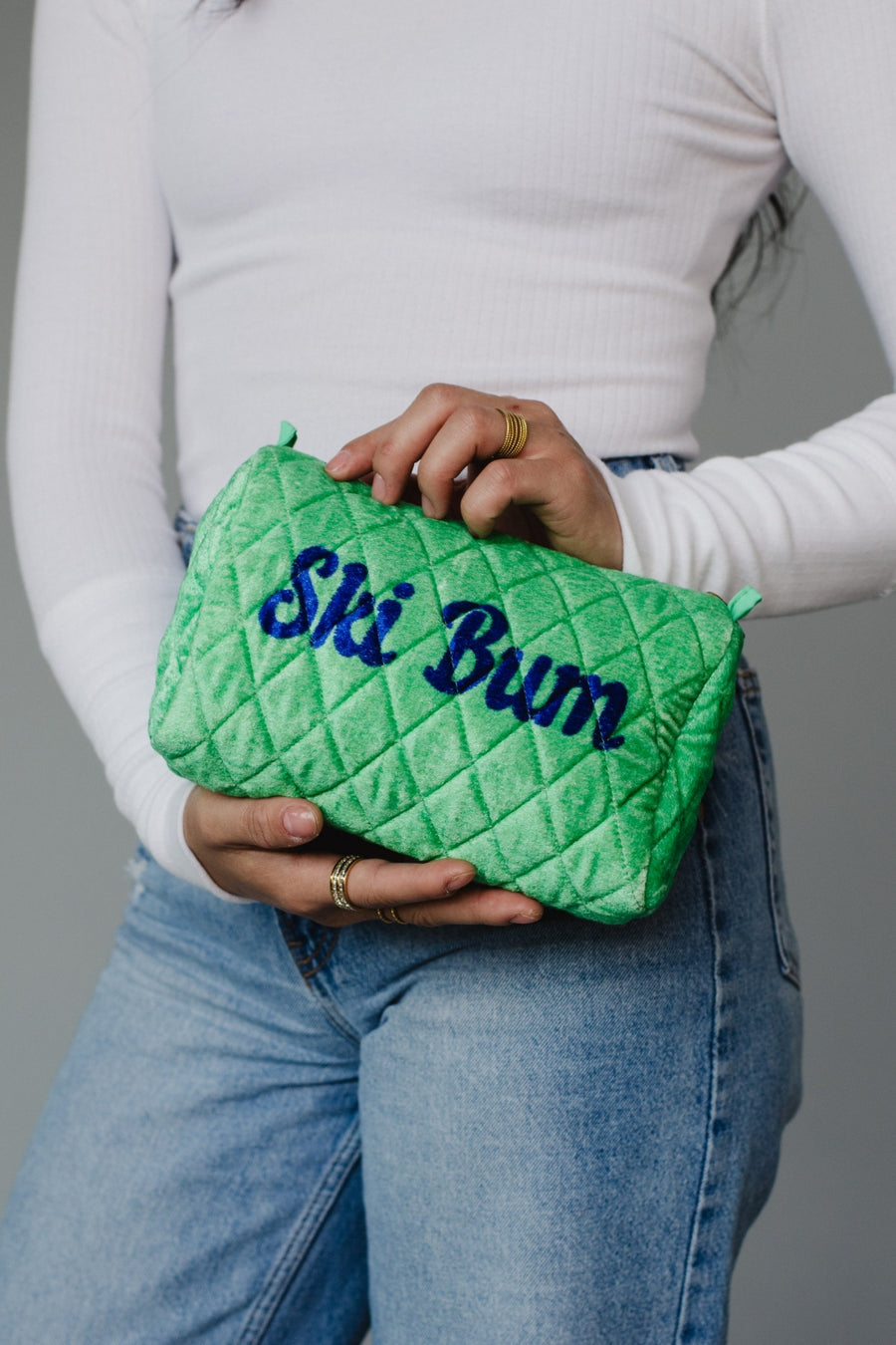Person holding a green quilted pouch with 'Ski Bum' text against a gray background