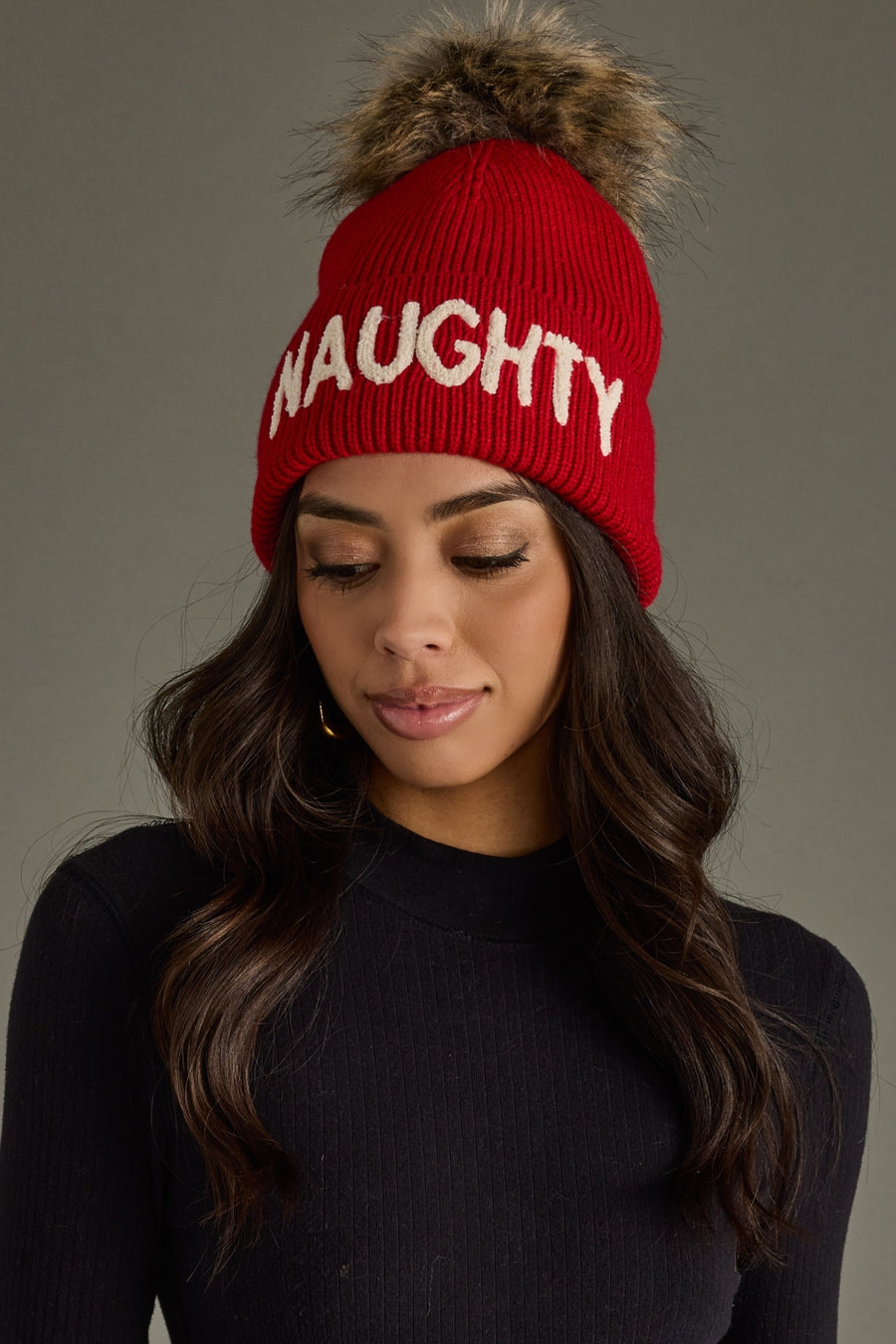 Woman wearing a red beanie with 'NAUGHTY' text and a fur pom-pom on a gray background