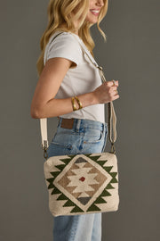 A woven crossbody bag features a striking geometric southwestern pattern with deep green triangular motifs and burgundy diamond accents on a natural beige textured base. The bag includes an adjustable crossbody strap with antique brass hardware, showcasing artisan craftsmanship with a bohemian-chic aesthetic against a rich teal background.