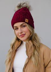Woman wearing a maroon knit beanie with a fur pom-pom against a gray background