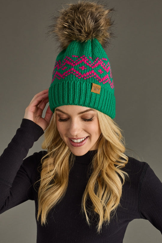 Woman wearing a green knit beanie with a pink pattern and fur pom-pom against a gray background